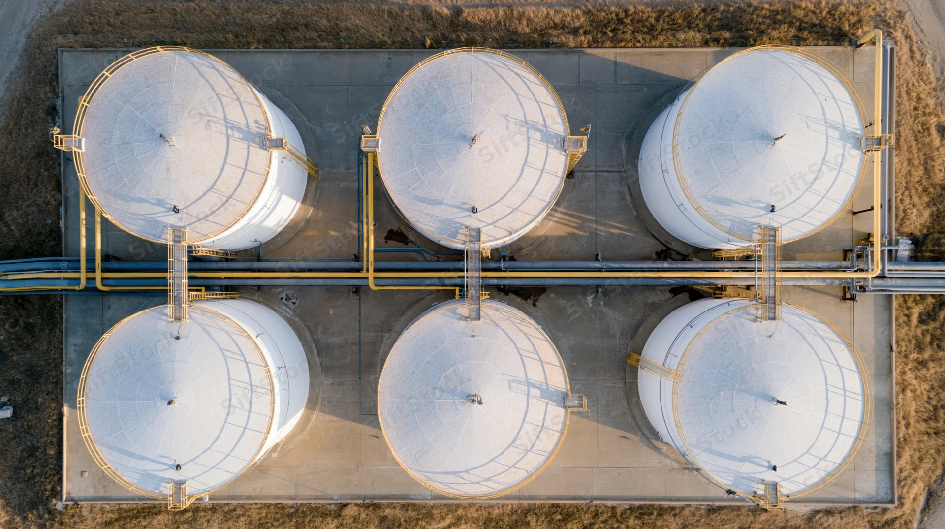 Aerial top-down view of six large white storage tanks arranged in two rows on a concrete foundation with connecting pipelines and access ladders