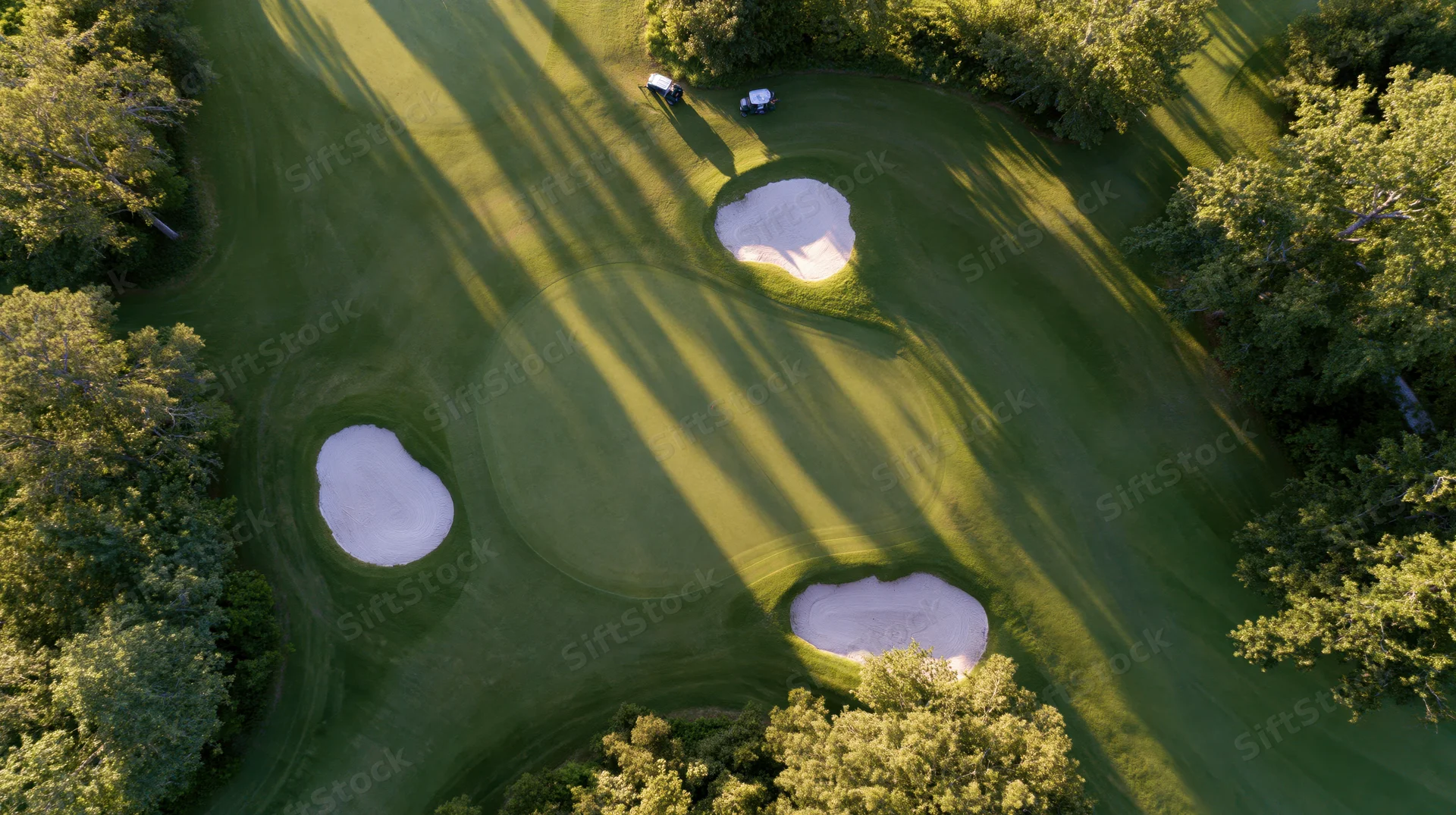 Aerial view of a golf course green with sand traps and trees surrounded by mowed grass and sunlight casting long shadows