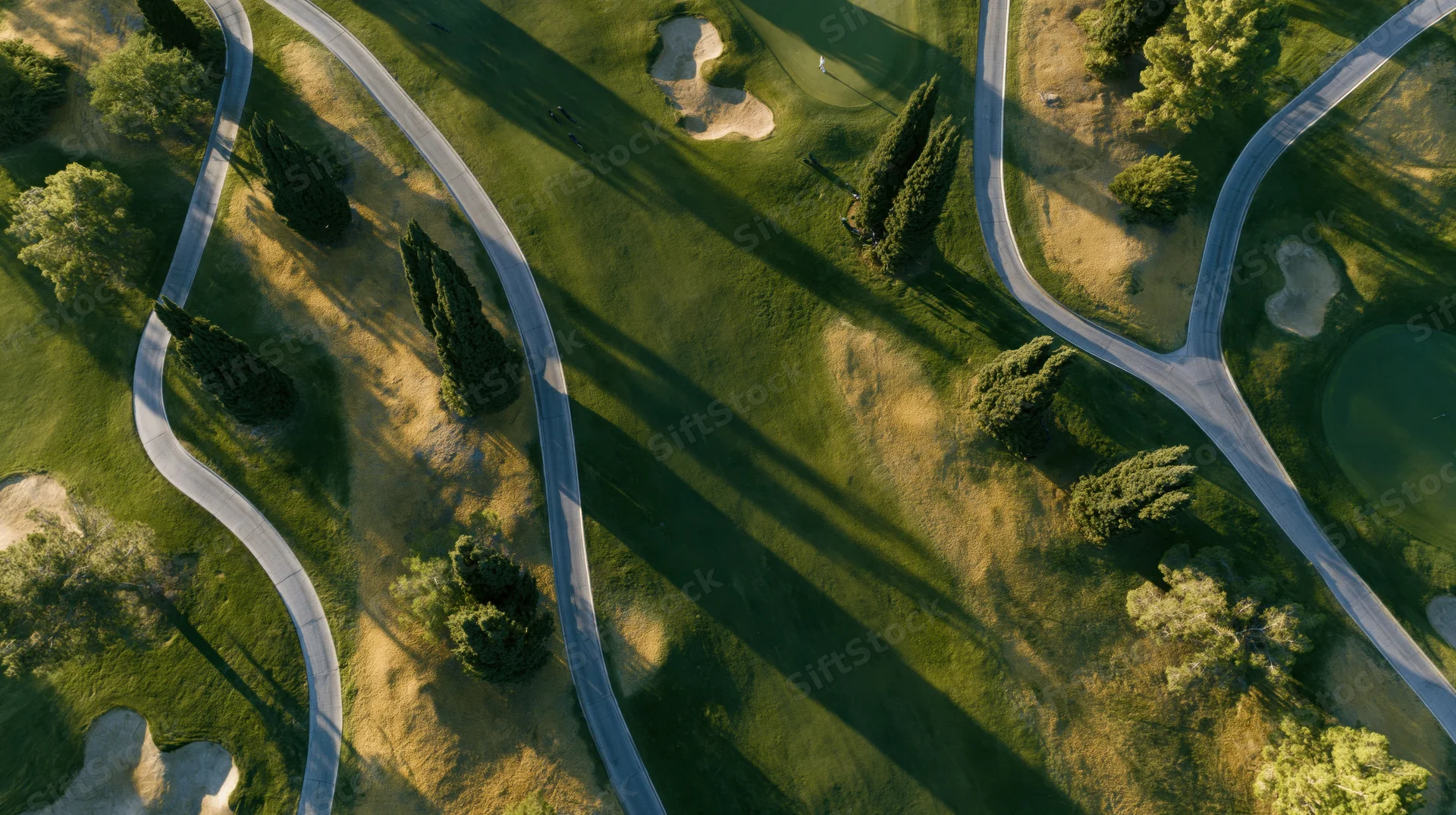 Aerial top-down view of a manicured golf course with winding paths, sand bunkers, and trees casting long shadows in sunlight