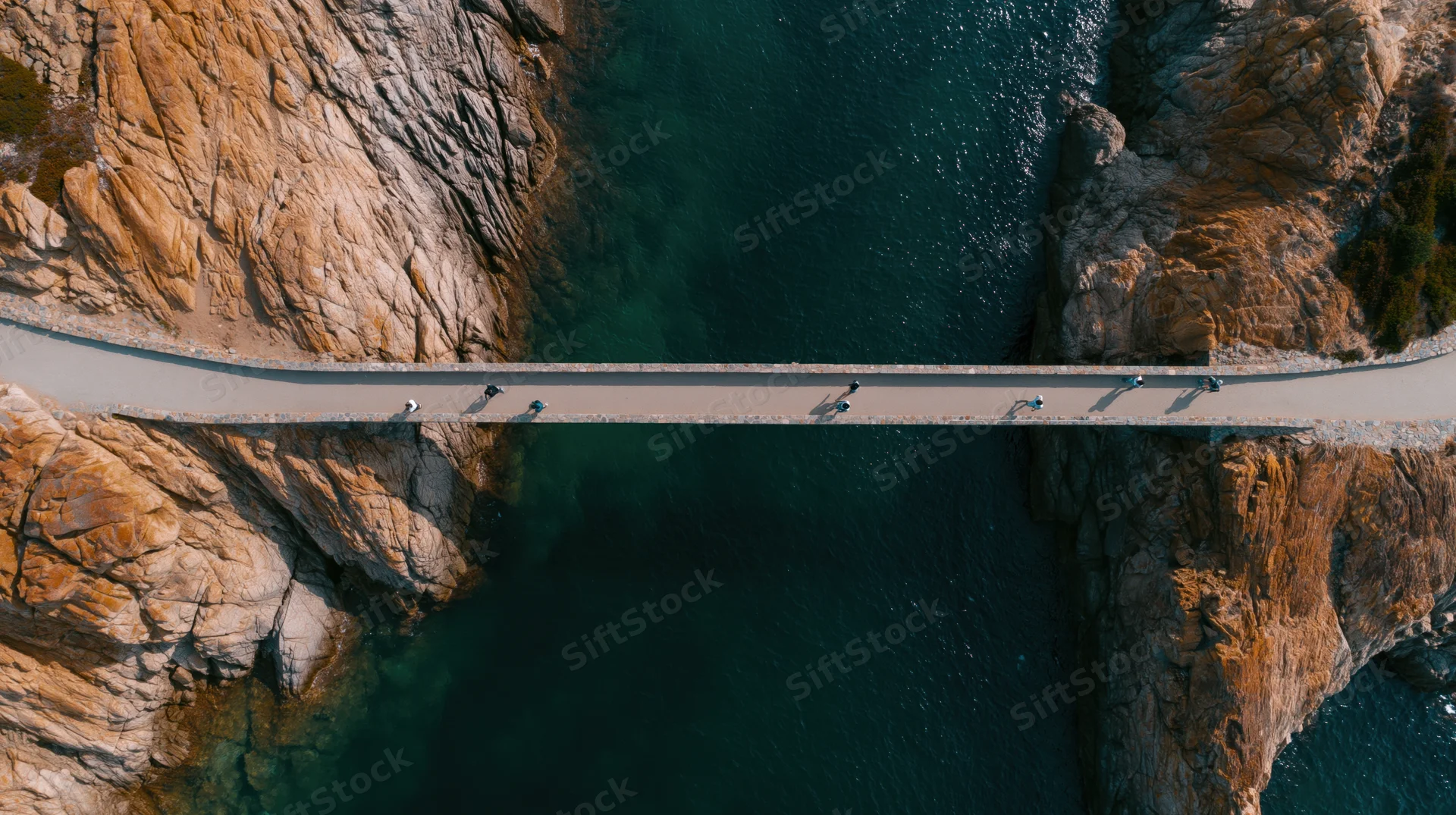 Aerial top-down view of narrow concrete pedestrian bridge over deep blue water between rocky cliffs with people walking across