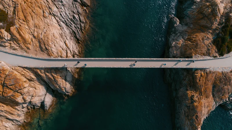 Aerial top-down view of narrow concrete pedestrian bridge over deep blue water between rocky cliffs with people walking across