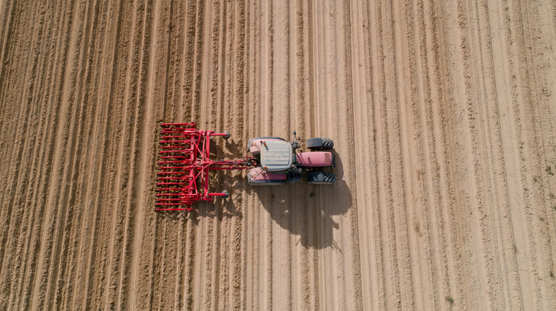 Pink and grey tractor pulling red plow across tilled field from above