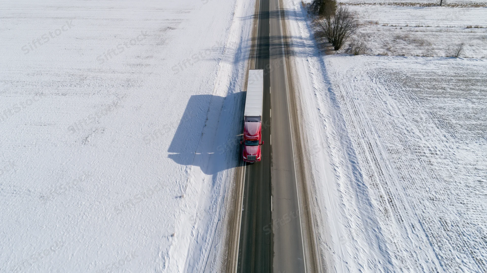Red semi-truck driving on snow-covered highway through rural winter landscape