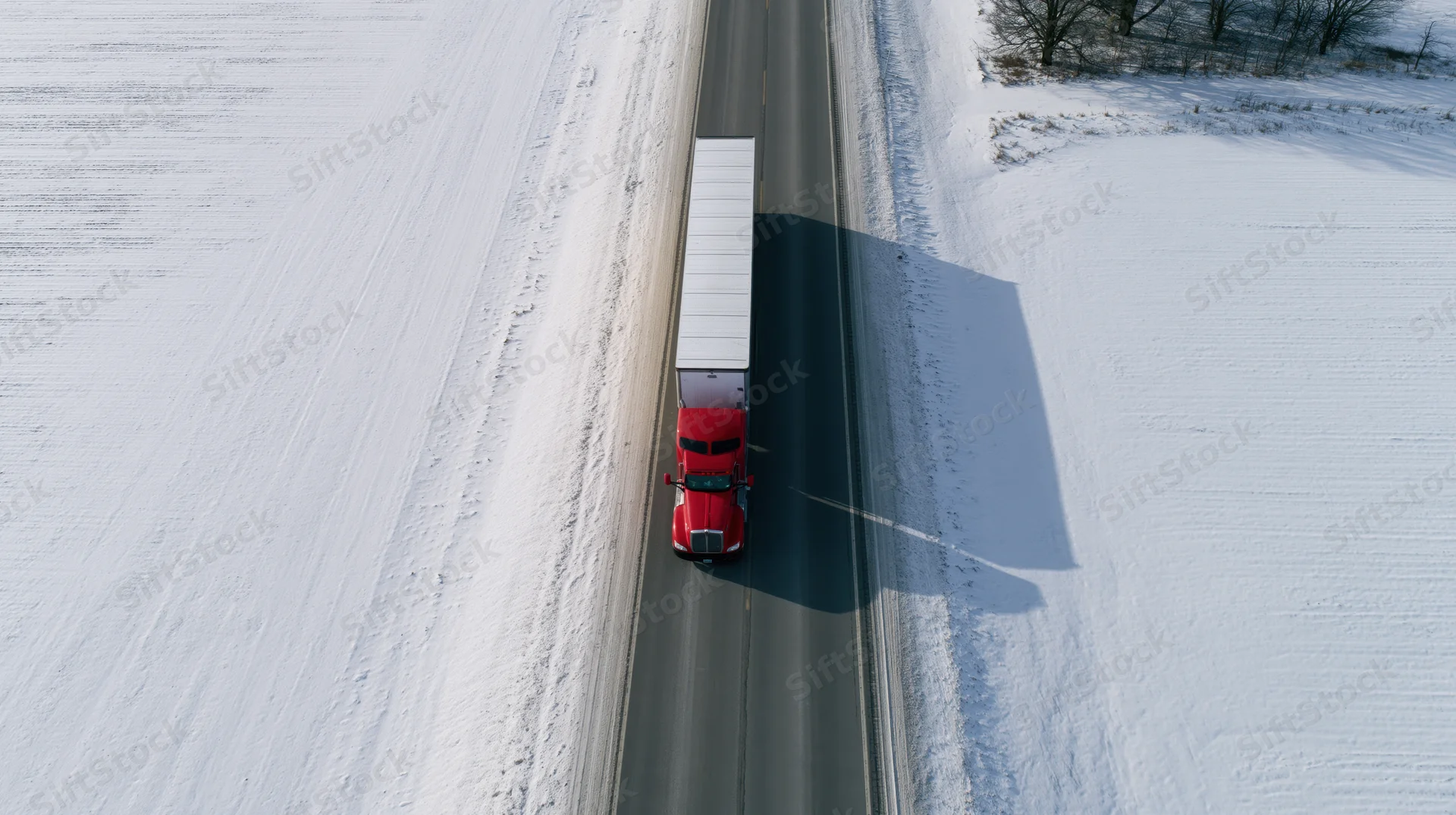 Red semi-truck with white trailer driving on snow-covered highway from aerial top-down view
