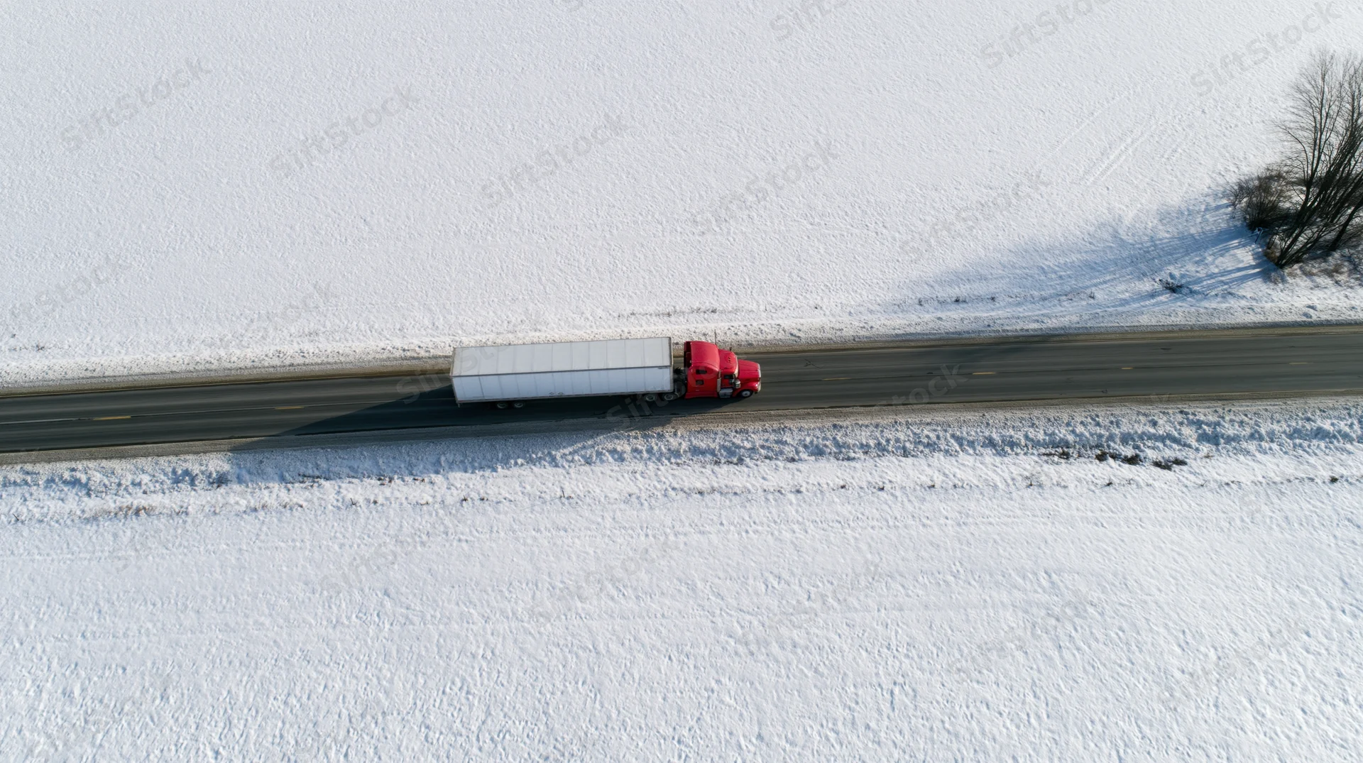 Red semi-truck with white trailer driving on snow-covered highway from aerial view