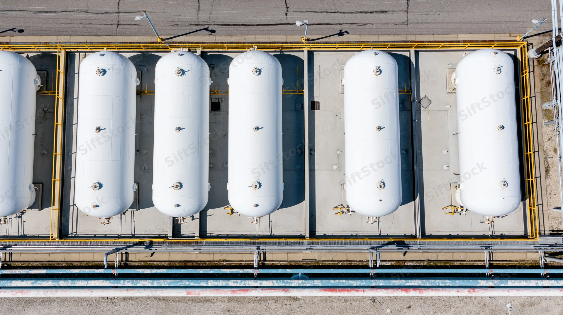 Aerial top-down view of six white cylindrical storage tanks arranged in a row on an industrial facility rooftop with yellow safety railings and concrete pavement