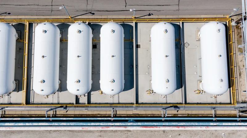 Aerial top-down view of six white cylindrical storage tanks arranged in a row on an industrial facility rooftop with yellow safety railings and concrete pavement