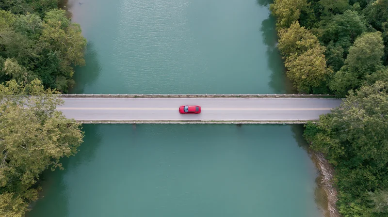 Red car driving across a bridge over a calm river surrounded by trees, aerial top-down view