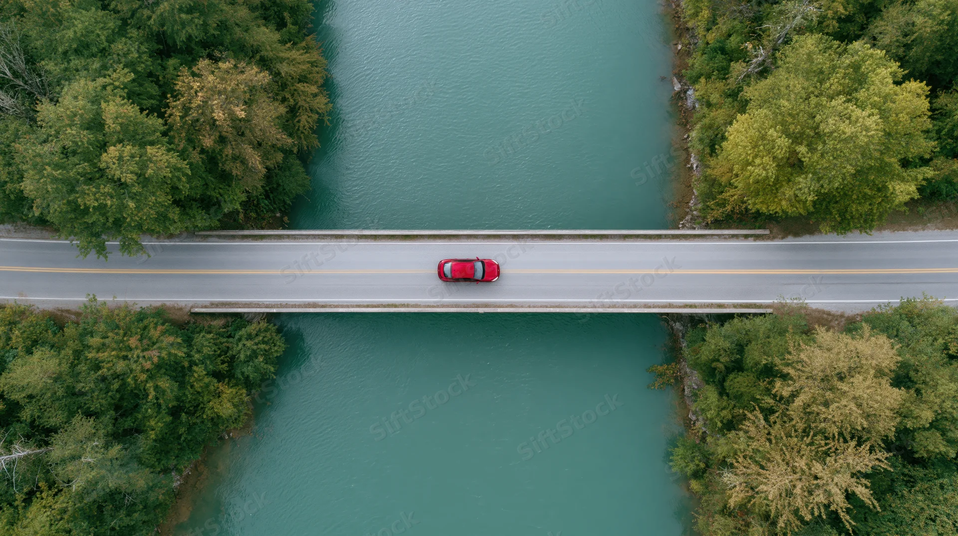 Red car driving across a bridge over a turquoise river surrounded by forest from aerial top-down view
