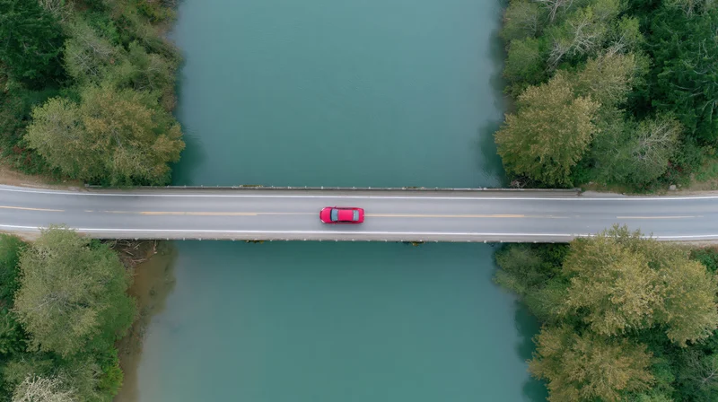 Red car driving across a bridge over a river from an aerial top-down view
