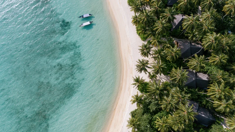 Aerial top-down view of a tropical beach resort with white sand, turquoise water, palm trees, and thatched-roof villas