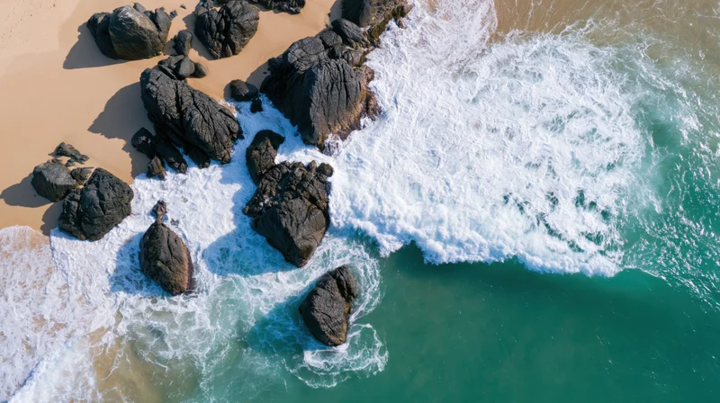 Aerial top-down view of ocean waves crashing over dark rocks along a sandy beach