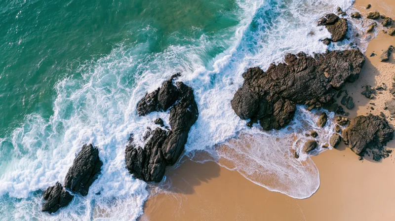 Aerial top-down view of ocean waves crashing over dark rocks along sandy beach