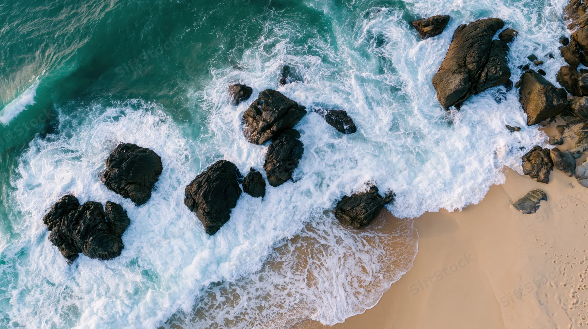 Aerial top-down view of ocean waves crashing over dark rocks along sandy beach
