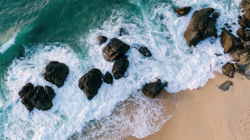 Aerial top-down view of ocean waves crashing over dark rocks along sandy beach