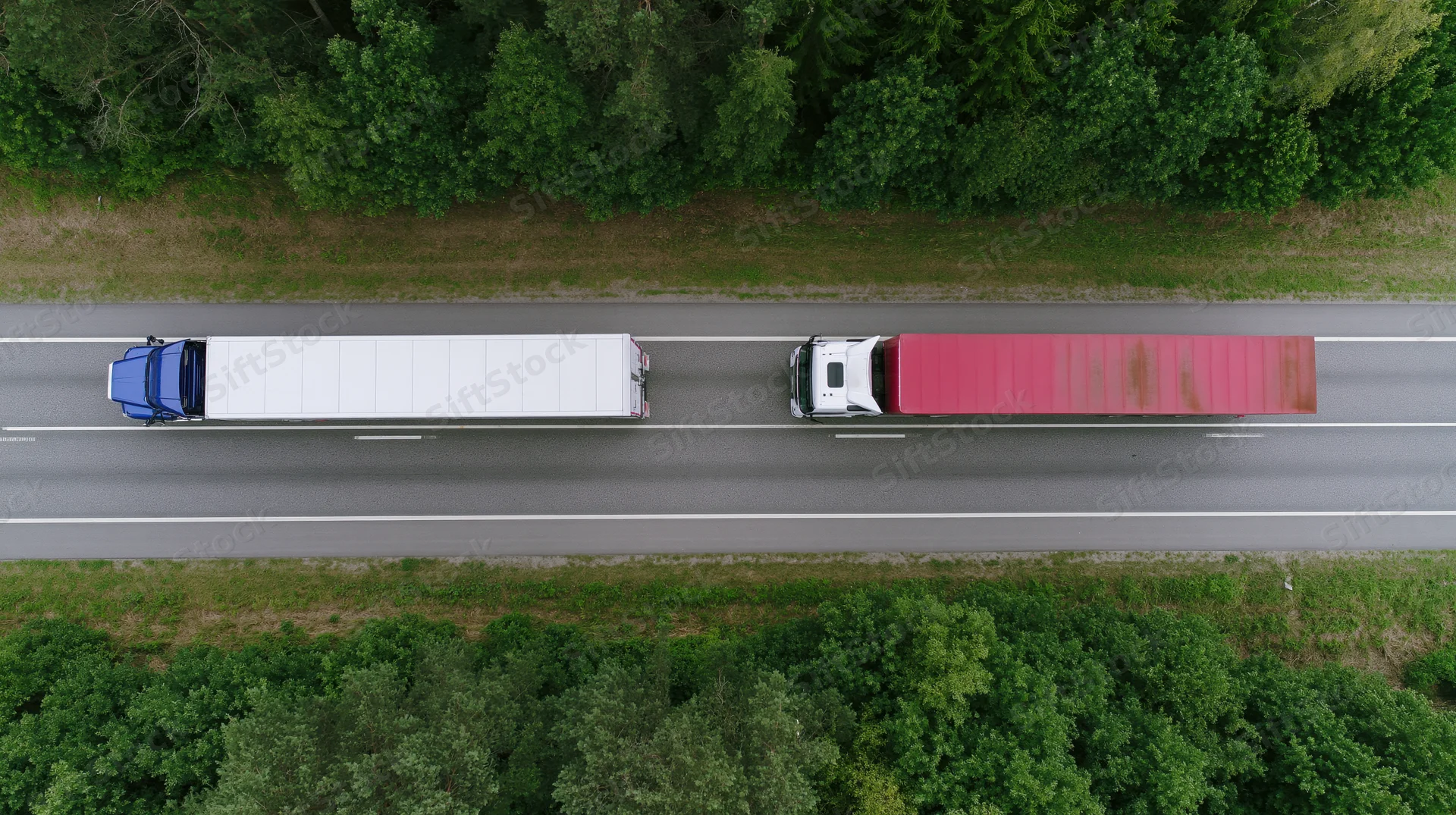 Two semi-trucks driving side by side on a highway surrounded by forest, aerial top-down view