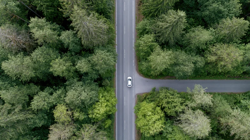 Aerial top-down view of a white car driving on a two-lane asphalt road through a dense green forest at an intersection