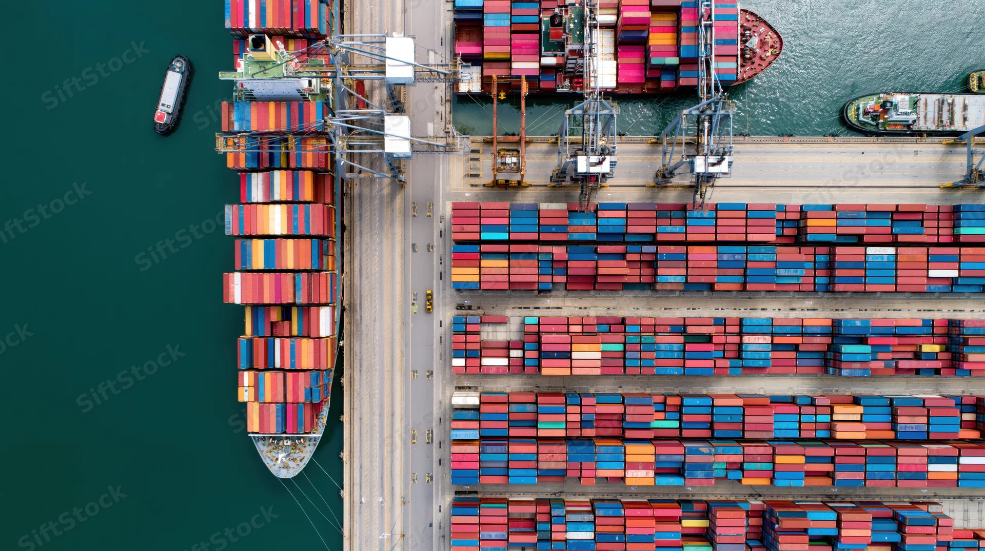 Aerial top-down view of a busy commercial shipping port with stacked containers and cranes at the dock