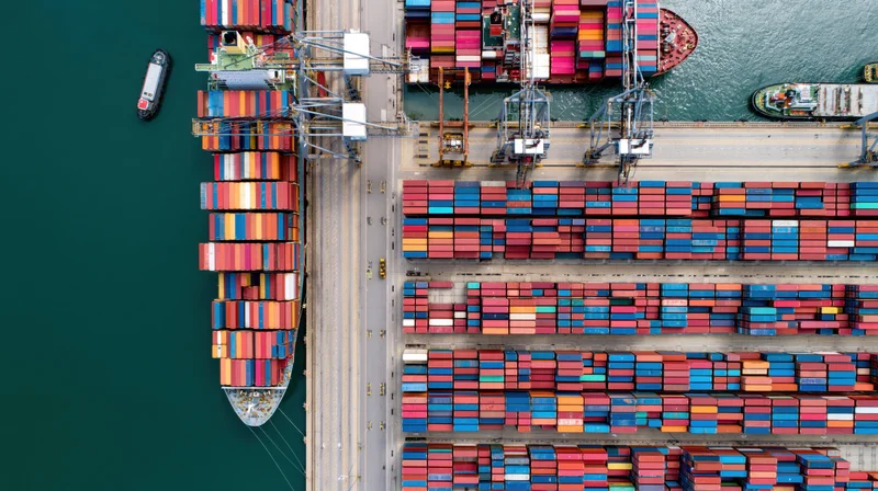 Aerial top-down view of a busy commercial shipping port with stacked containers and cranes at the dock