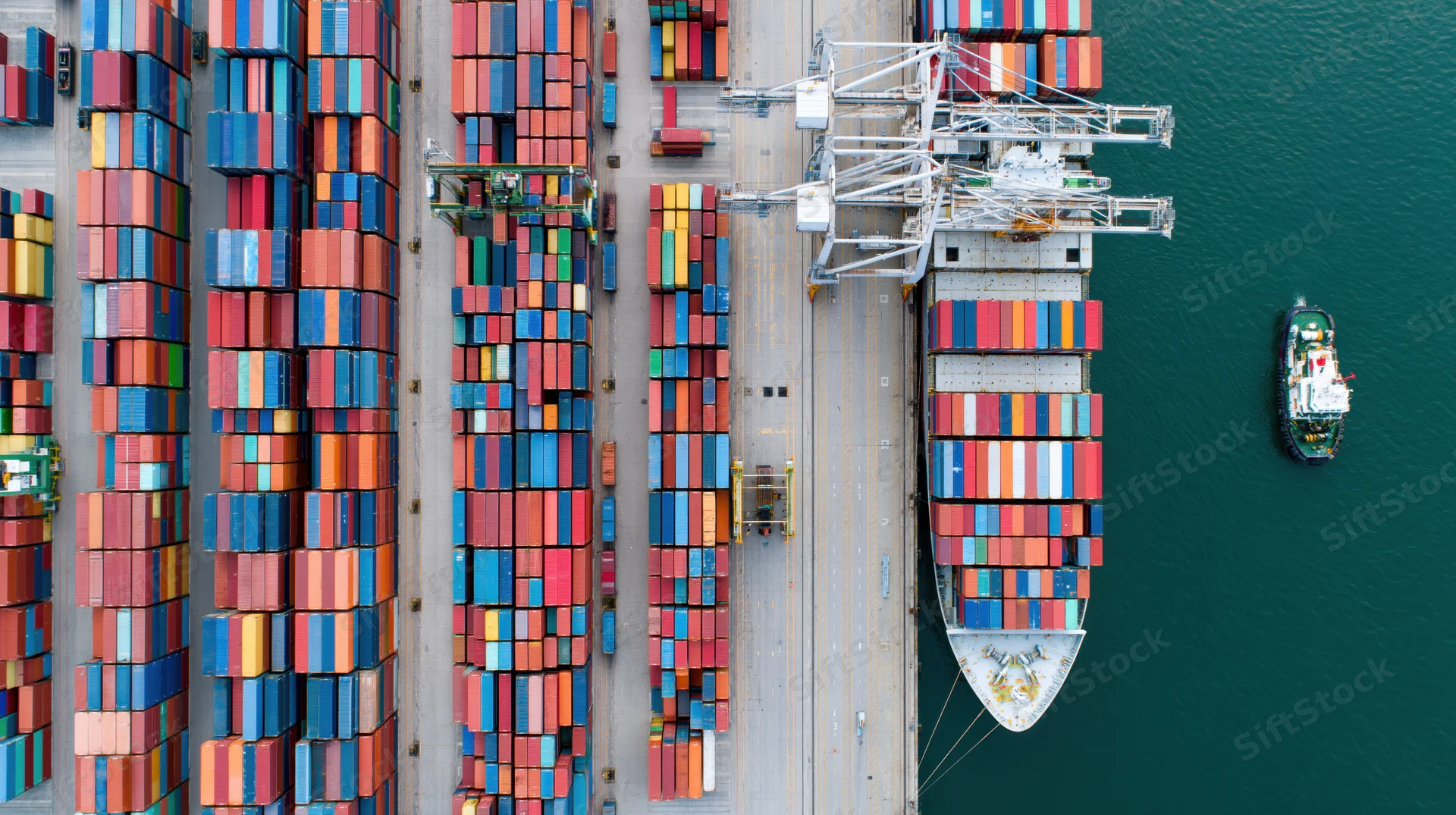 Aerial top-down view of a busy commercial shipping port with stacked containers, cranes, and a cargo ship docked at the terminal