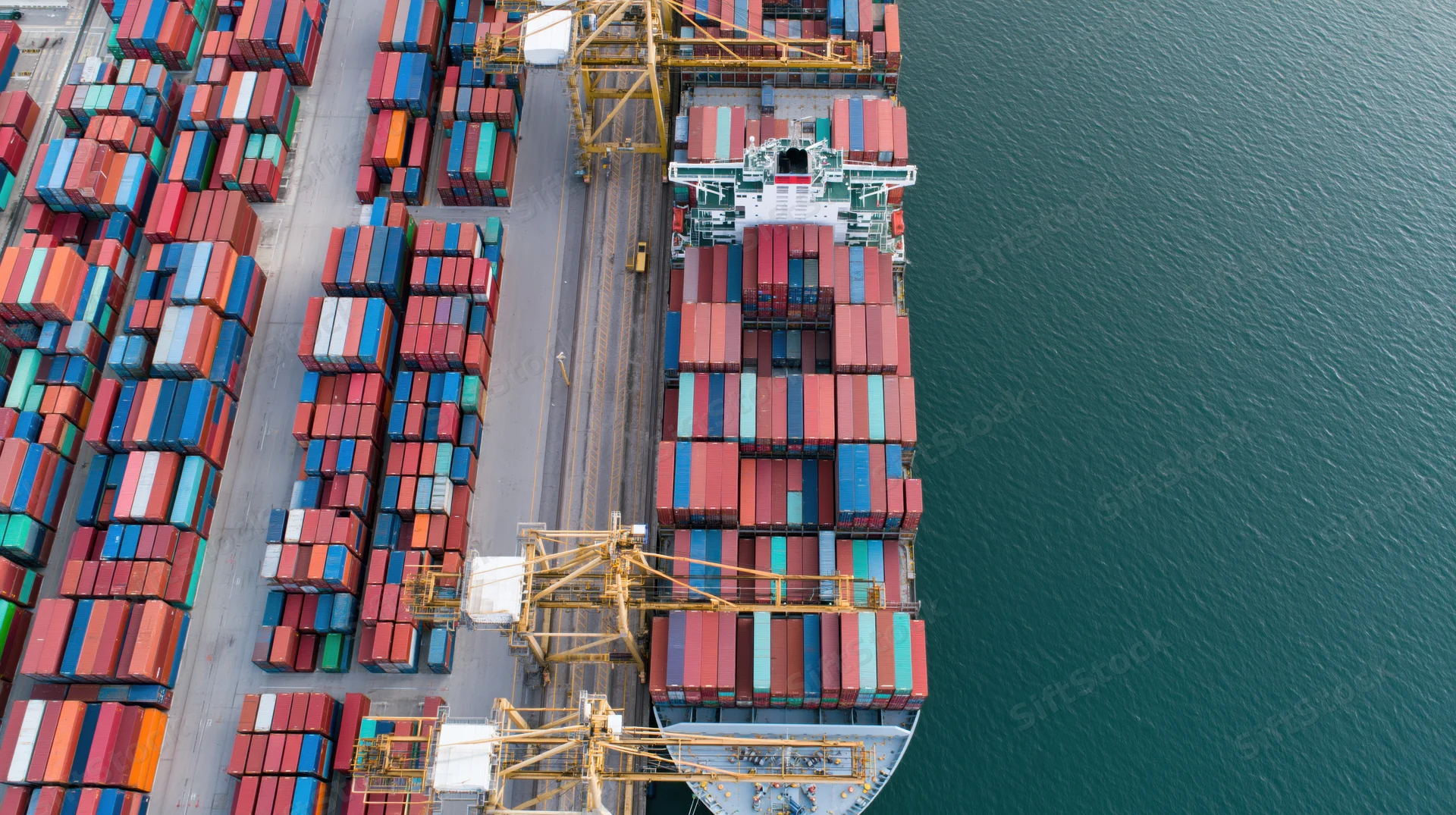 Aerial top-down view of a large container ship docked at a commercial port with stacked shipping containers and cranes loading cargo