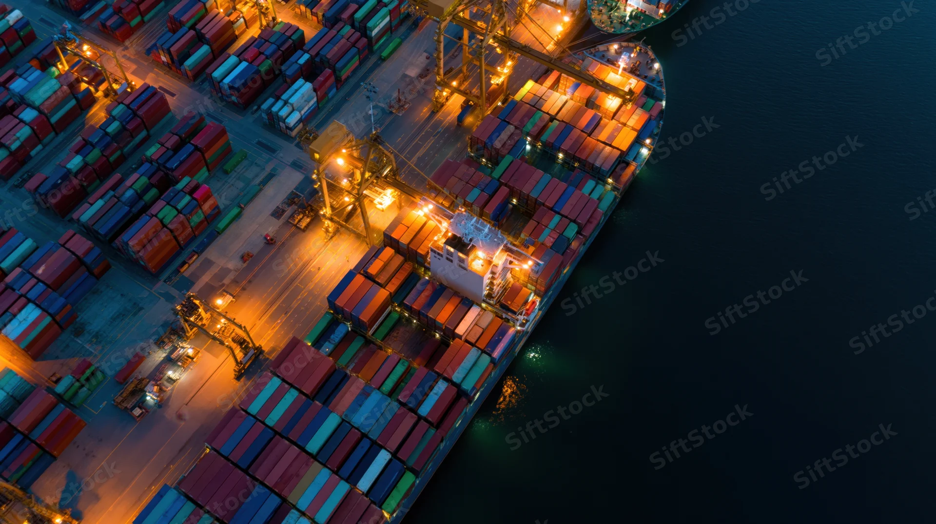 Large container ship docked at port terminal at night with cranes loading cargo under artificial lights