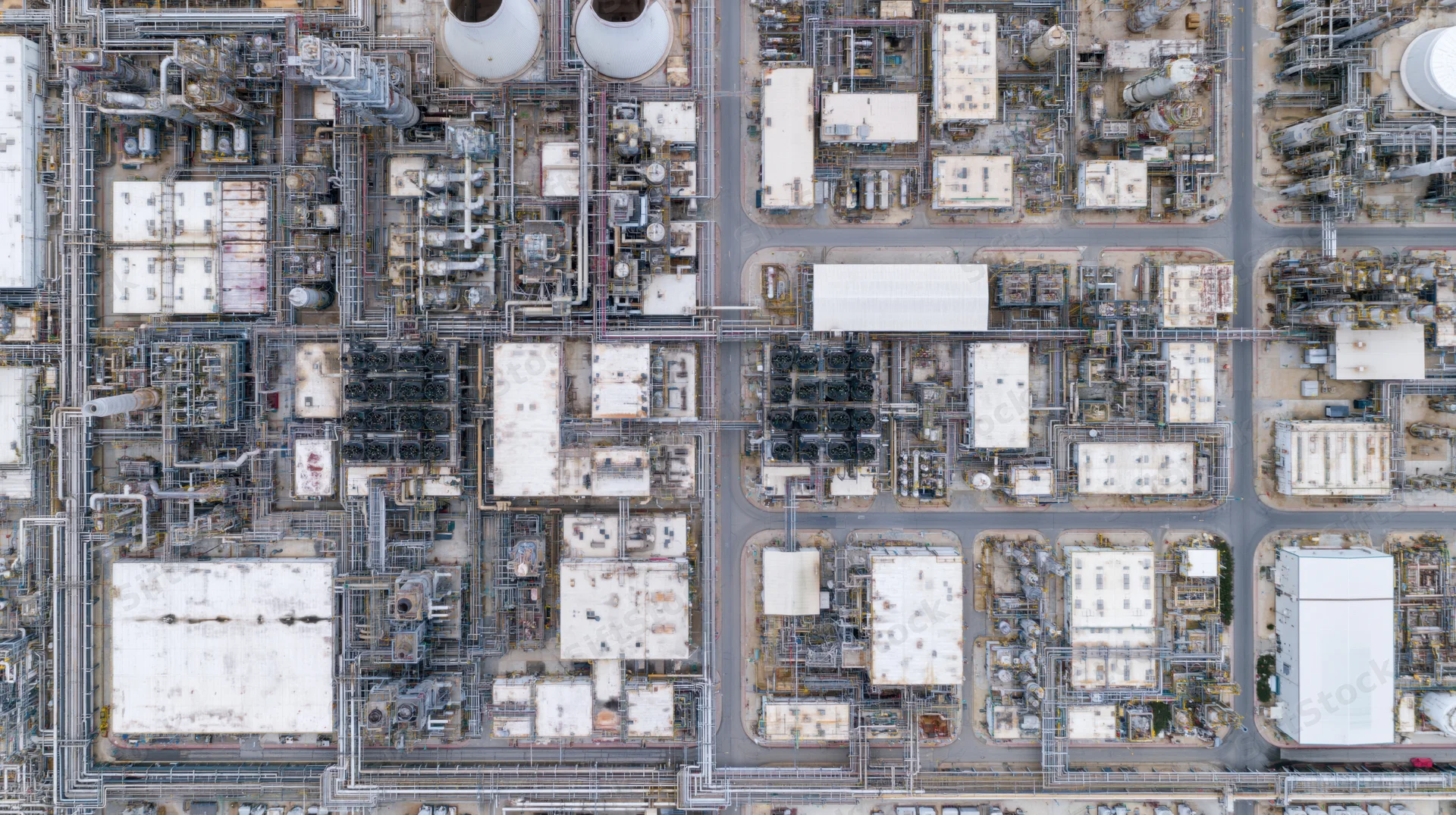 Aerial top-down view of a large petrochemical plant with interconnected pipes, storage tanks, and processing units arranged in a grid pattern across an industrial complex