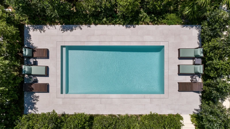 Aerial top-down view of a rectangular outdoor swimming pool surrounded by lounge chairs and green trees on a tiled patio
