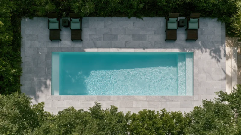 Aerial top-down view of a rectangular outdoor swimming pool surrounded by gray stone patio and green foliage with lounge chairs arranged along the edges