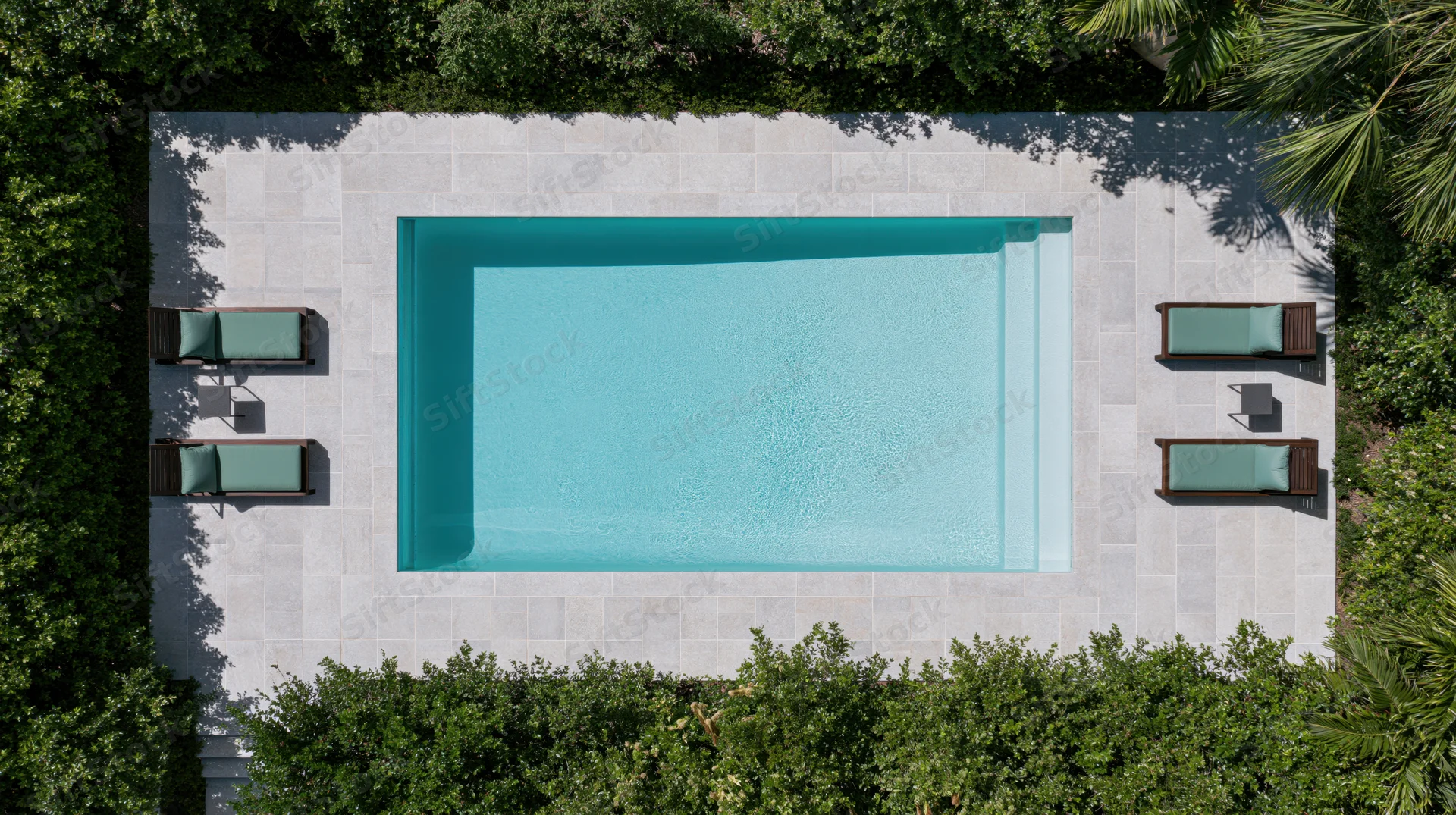 Aerial top-down view of a rectangular outdoor swimming pool surrounded by lounge chairs and green foliage on a stone patio