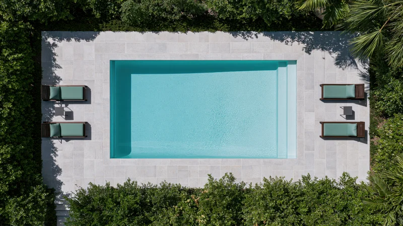 Aerial top-down view of a rectangular outdoor swimming pool surrounded by lounge chairs and green foliage on a stone patio