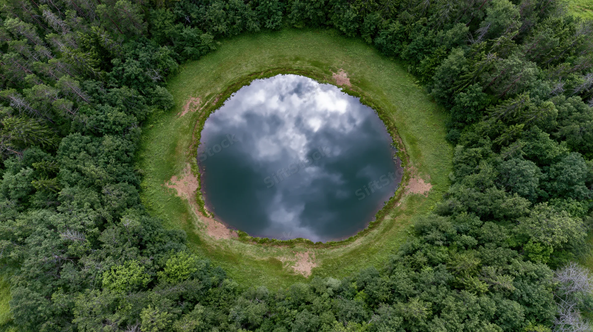 Aerial top-down view of a circular lake surrounded by dense forest, reflecting cloudy sky