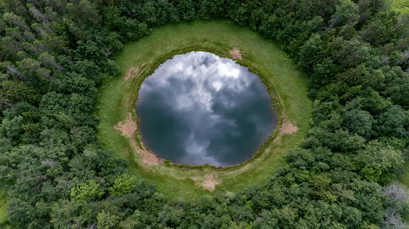 Aerial top-down view of a circular lake surrounded by dense forest, reflecting cloudy sky