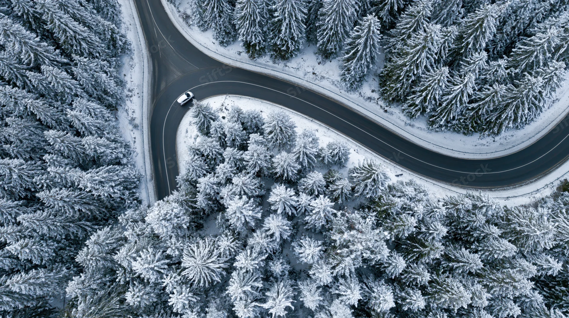 Aerial view of a winding road through a snow-covered pine forest with a car driving along it