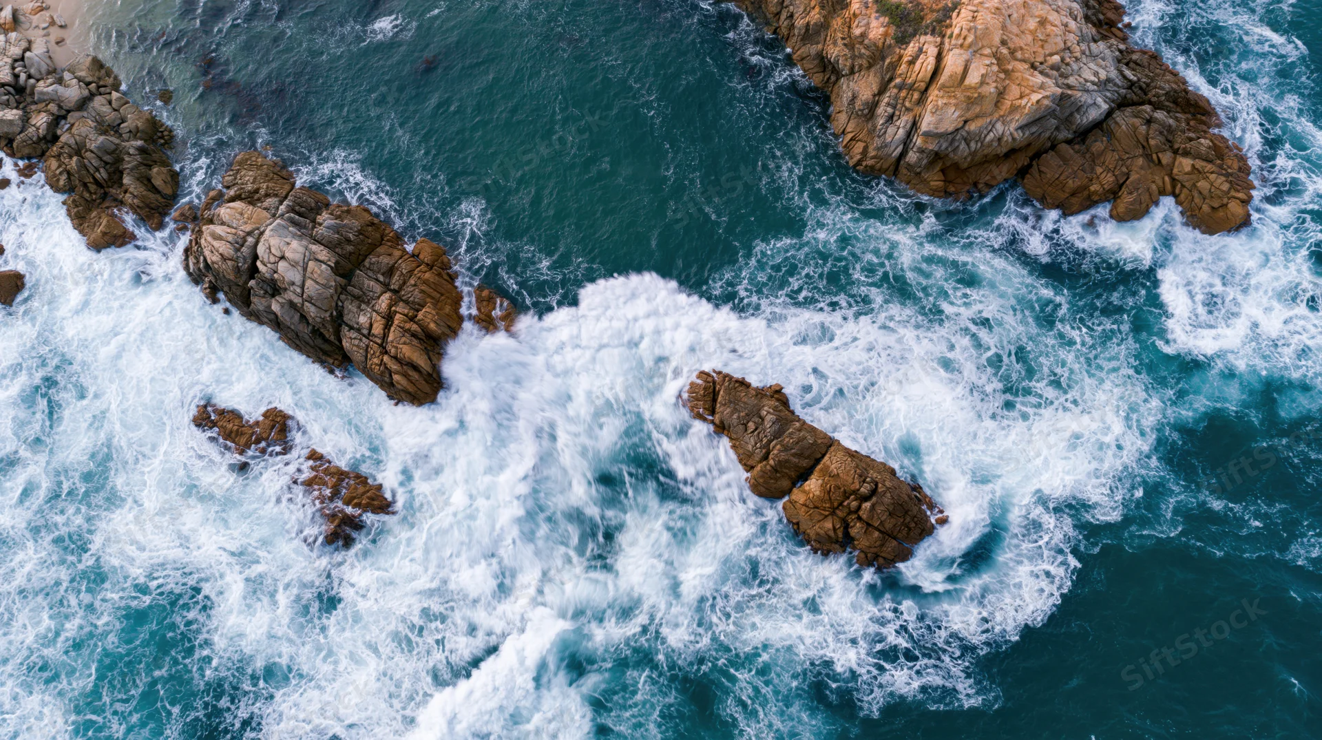 Aerial top-down view of ocean waves crashing against rugged rocks along the coastline