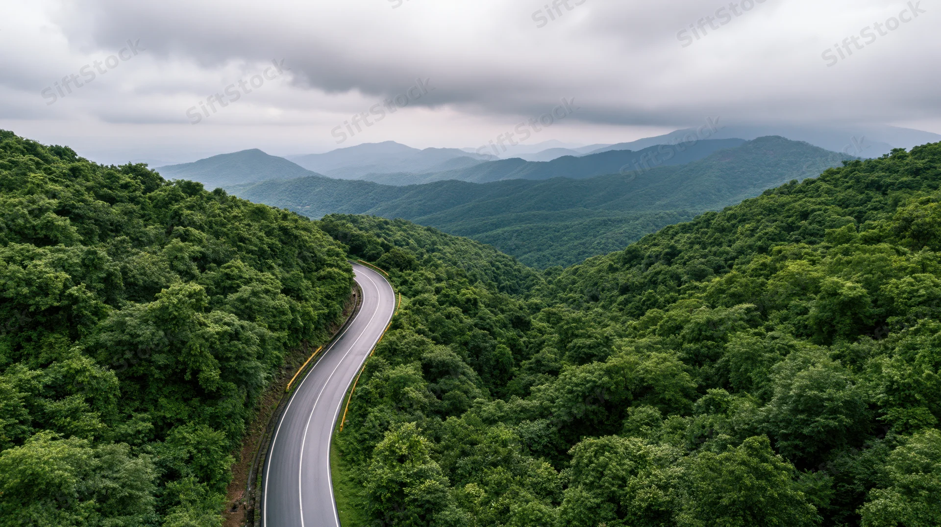 Winding asphalt road curving through dense green forested mountains under cloudy sky