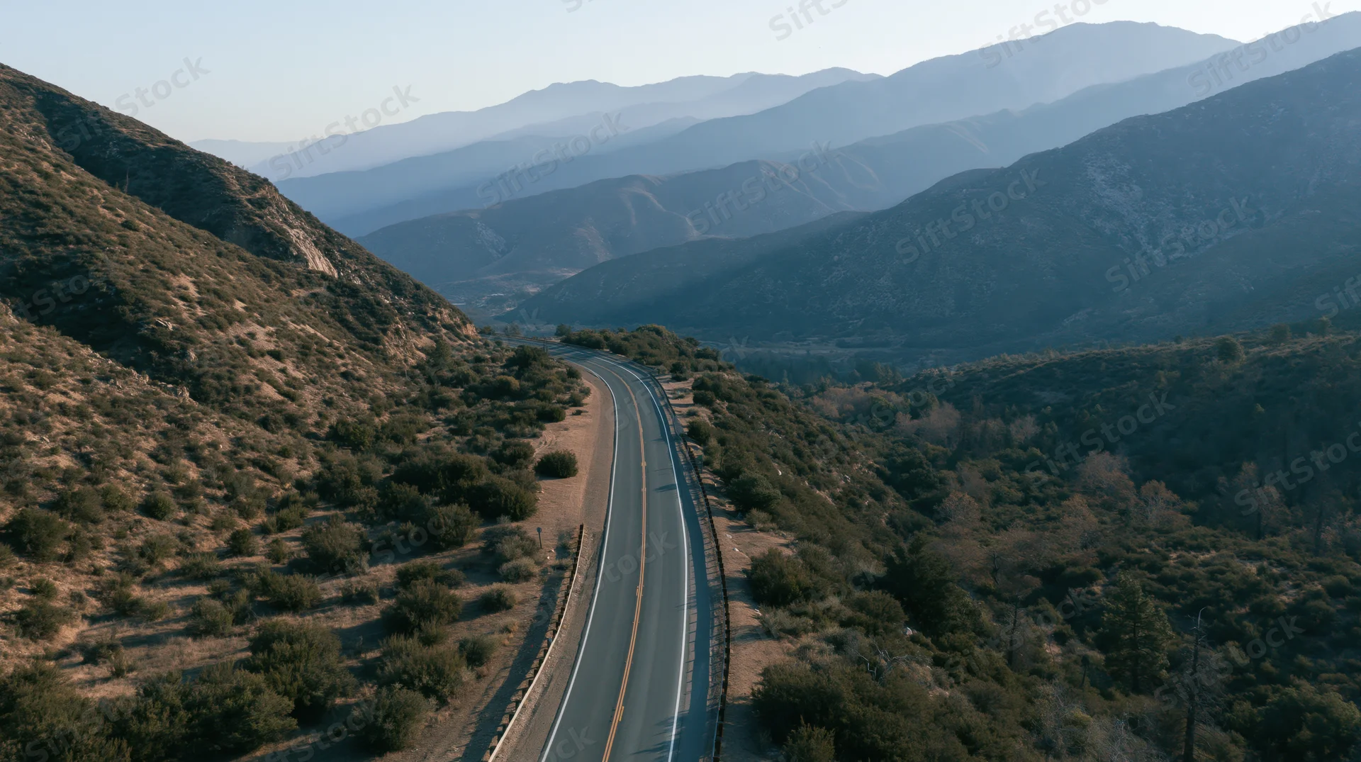 Aerial view of a winding two-lane highway through mountainous terrain with sparse vegetation and layered ridges in the distance