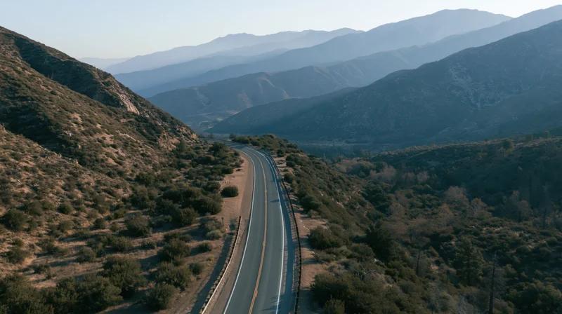 Aerial view of a winding two-lane highway through mountainous terrain with sparse vegetation and layered ridges in the distance