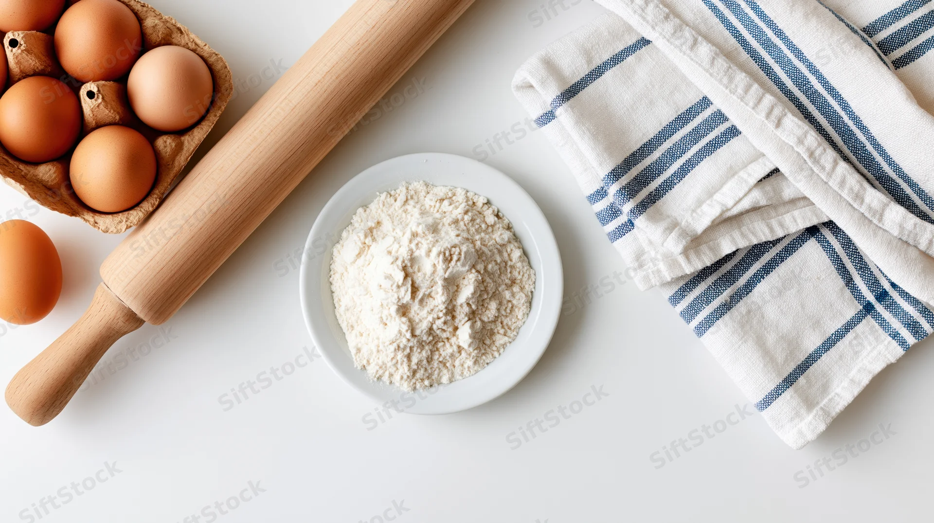 Baking ingredients including flour, eggs, and a rolling pin arranged on a white surface with a striped towel