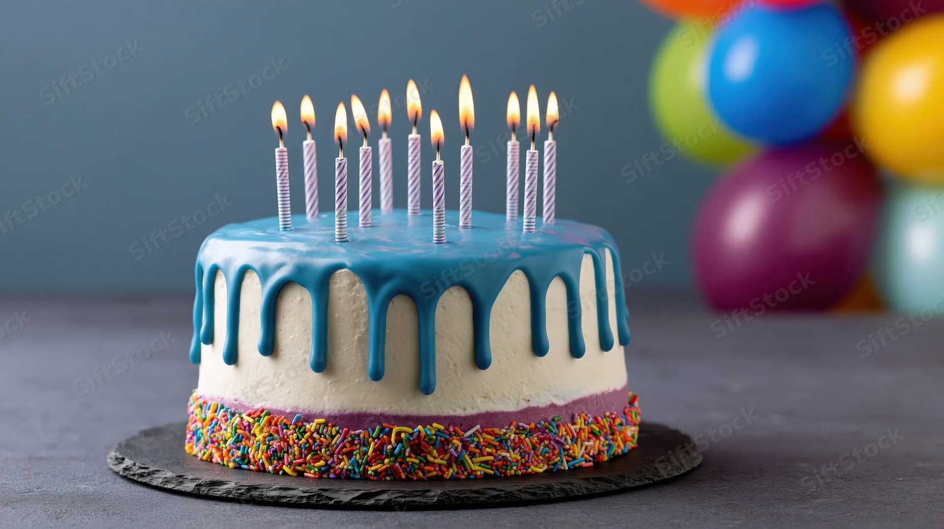 Birthday cake with lit candles and colorful sprinkles on dark slate surface, balloons in background