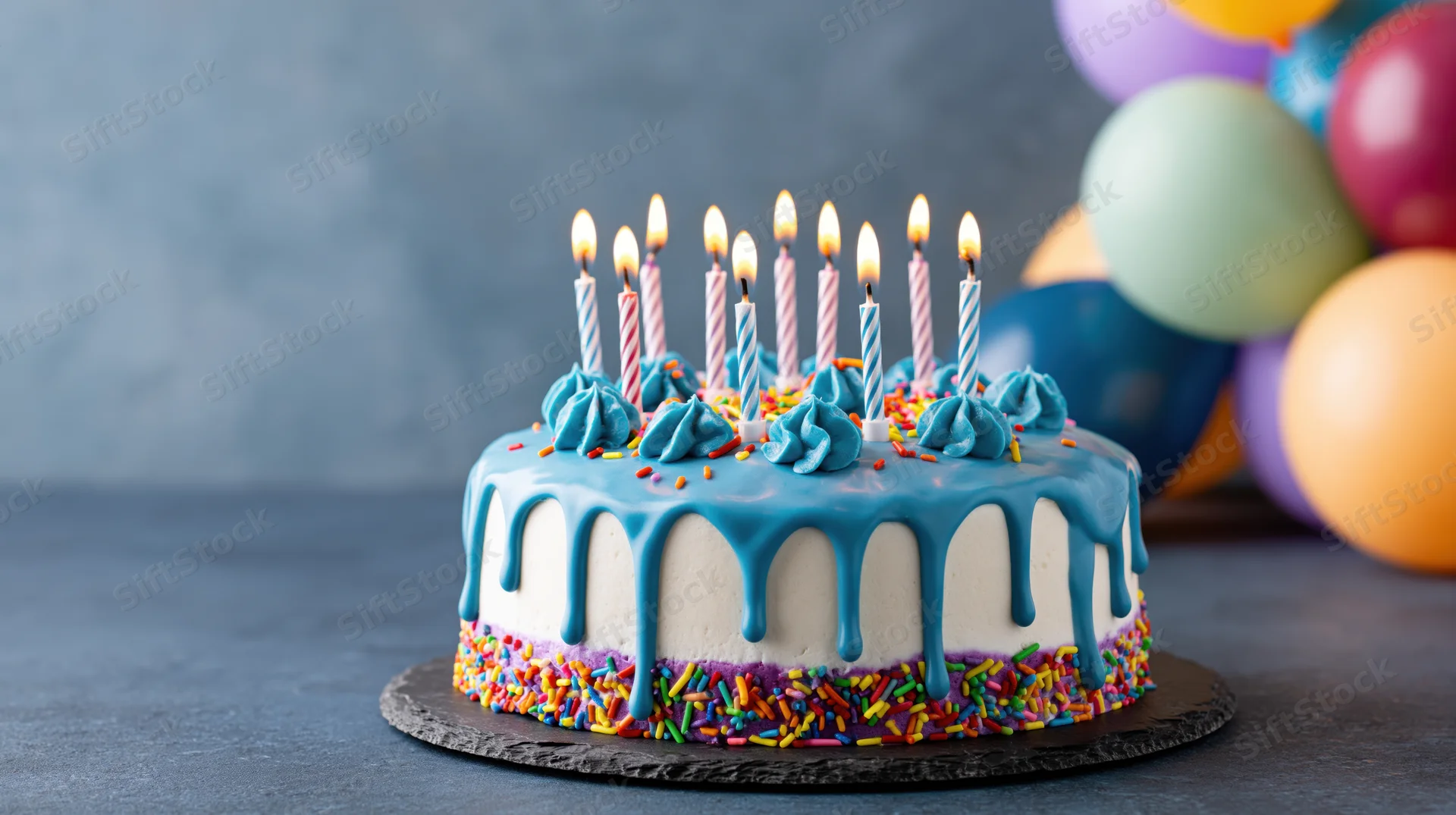 Birthday cake with lit candles and blue frosting drip on dark slate surface, colorful balloons in background