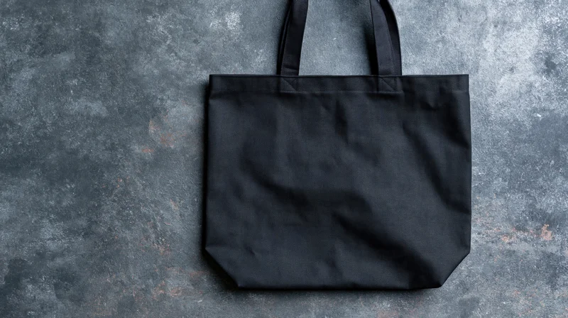 Black canvas tote bag lying flat on textured gray surface with handles upright