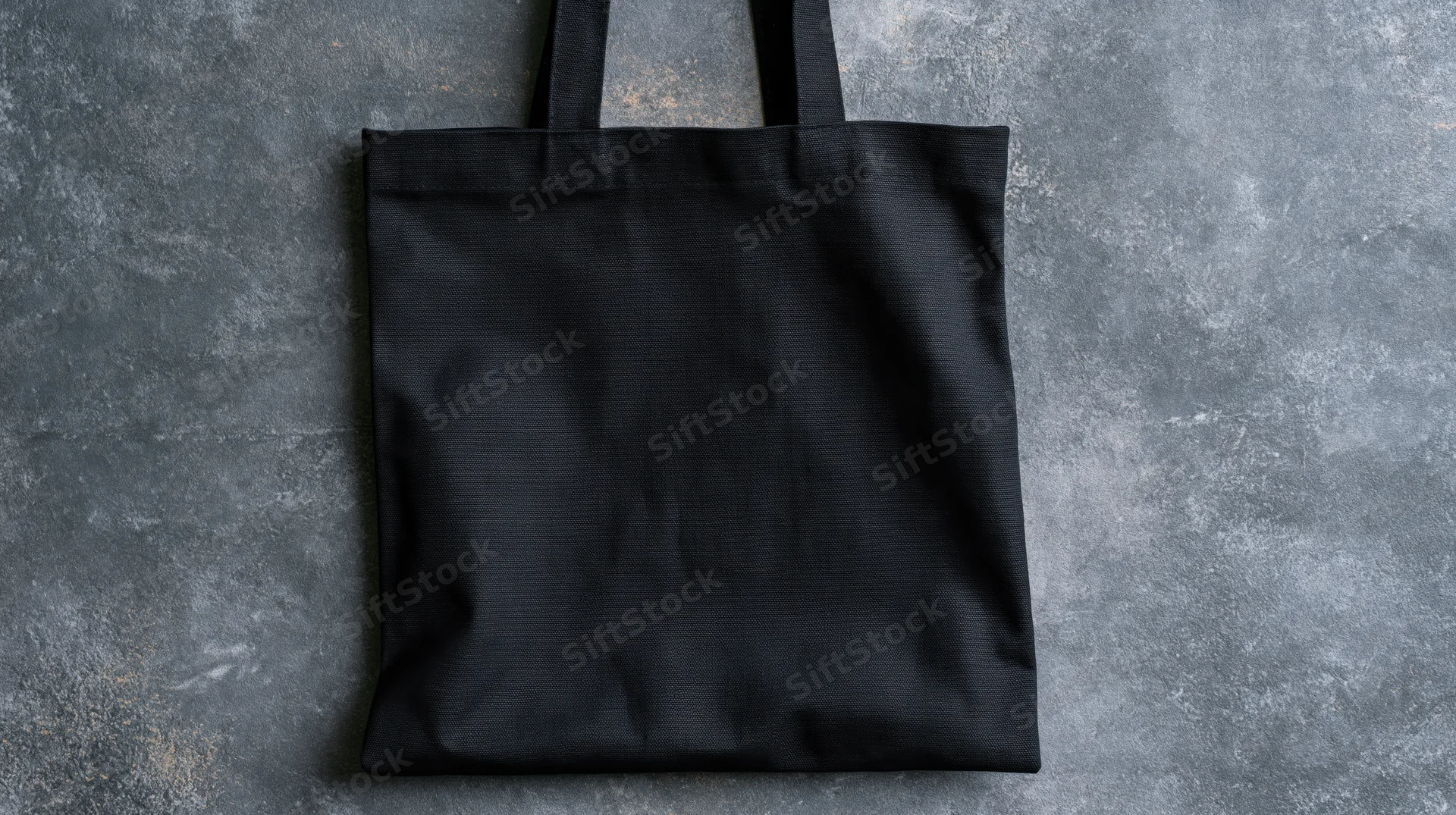 Black canvas tote bag lying flat on textured gray surface with handles upright