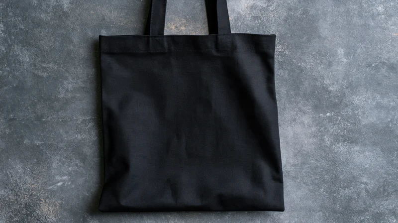 Black canvas tote bag lying flat on textured gray surface with handles upright