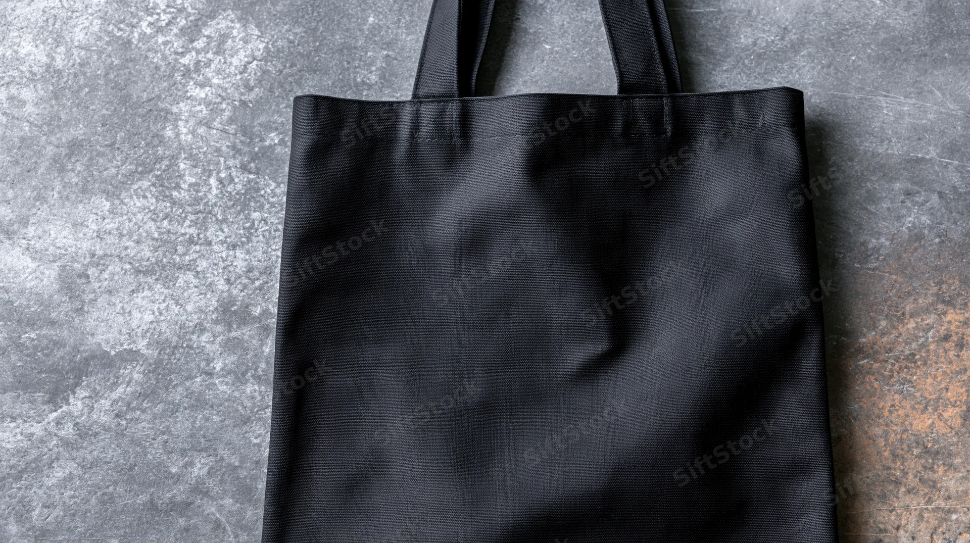 Black canvas tote bag lying flat on textured concrete surface with handles upright