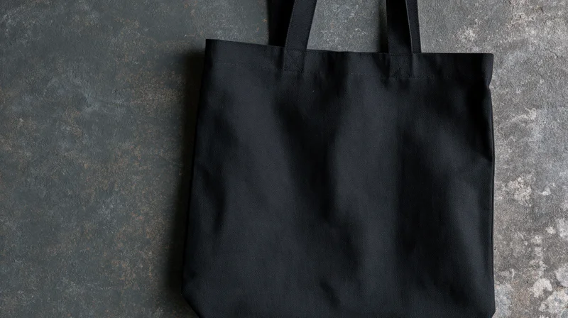 Black canvas tote bag lying flat on concrete surface with handles upright