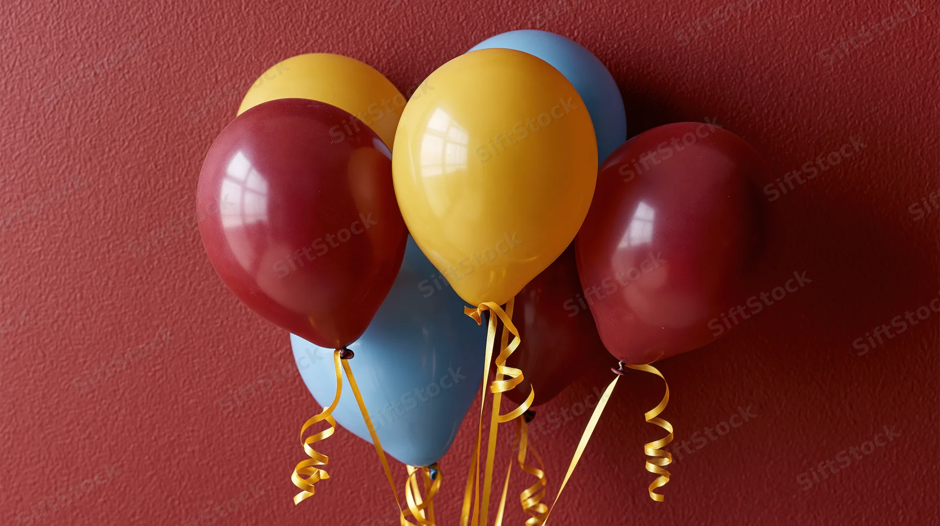 Cluster of burgundy yellow and blue latex balloons with gold ribbons against a red textured wall