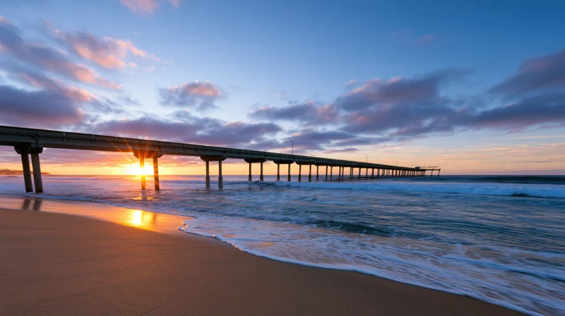 Long concrete fishing pier extending over ocean at sunset with waves washing onto sandy beach