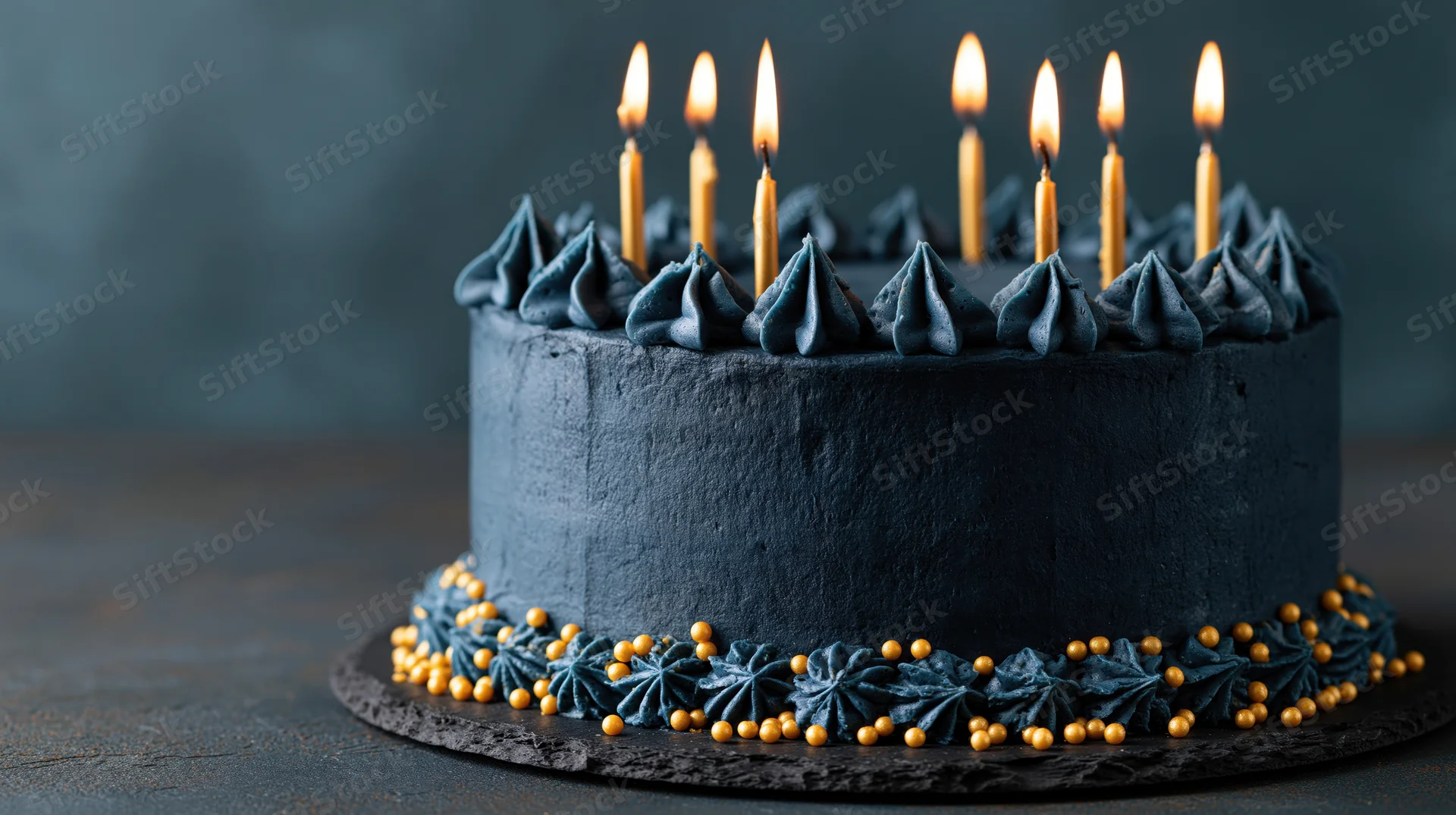 Dark navy birthday cake with six lit gold taper candles on a slate plate