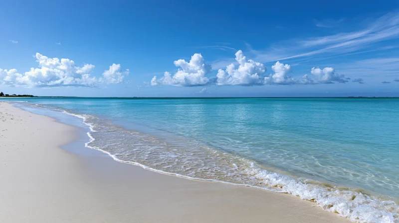 Calm turquoise water lapping onto white sand beach under blue sky with clouds